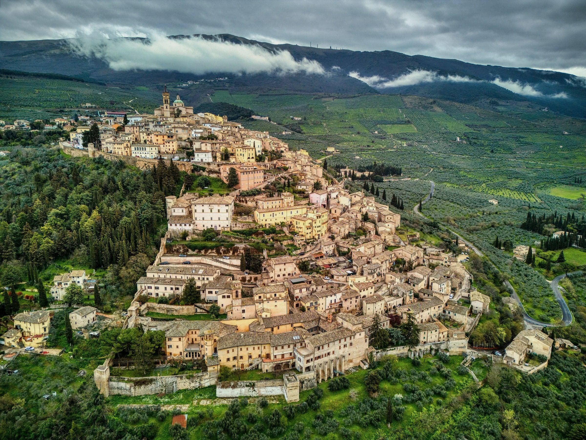 Trevi sul colle vista dall'alto, tra uliveti e boschi, con nuvole basse sulle montagne